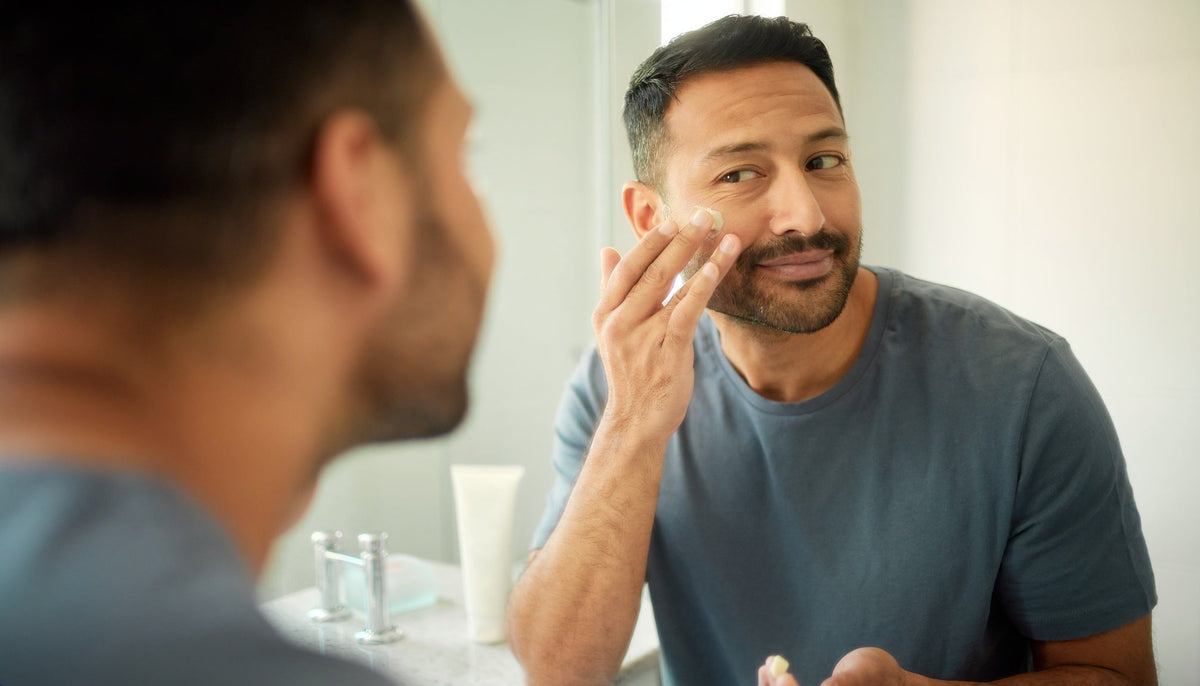 Adult man applying moisturiser to his cheek in a bright bathroom, with sunscreen in the background, framed above the navel.